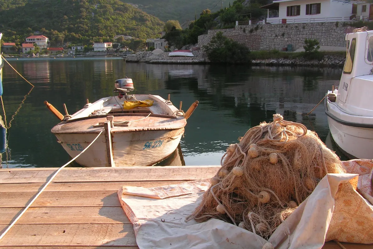 A wooden rowing boat and a fishing net on a jetty in Okuklje bay on Mljet, with the calm bay, the village houses and the wooded hills behind, in evening light