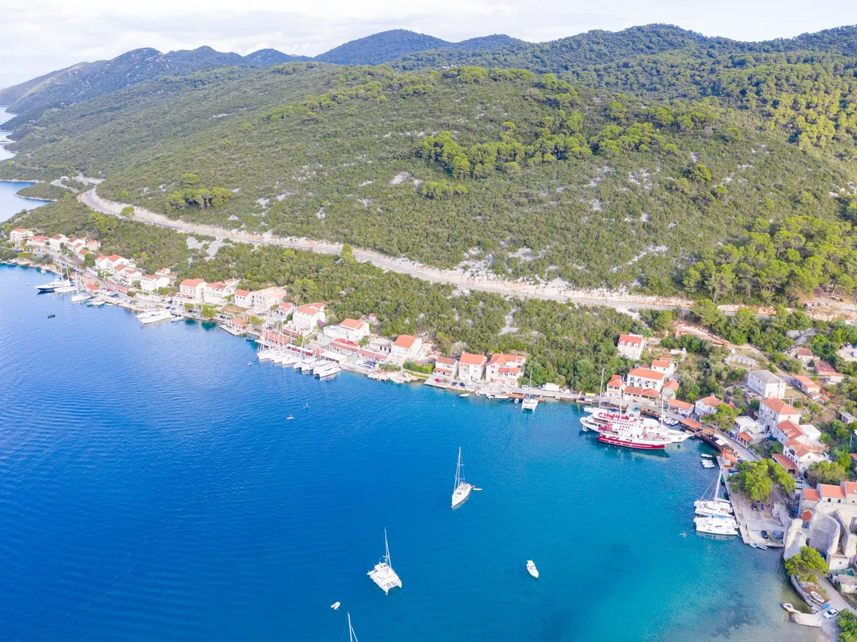 Polače on Mljet from the air, with yachts moored along the quay below the pine-covered hill, and sailing yachts and a catamaran at anchor in the bay