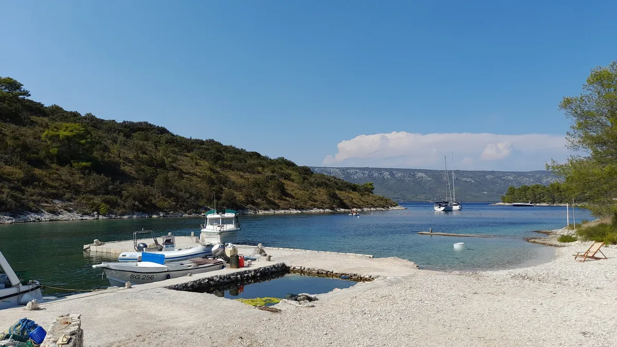 Mostir bay on Šćedro: small boats at a stone pier by the pebble beach, a yacht moored in the bay and the hills of Hvar across the channel