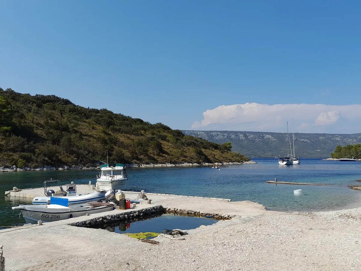 Mostir bay on Šćedro: small boats at a stone pier by the pebble beach, a yacht moored in the bay and the hills of Hvar across the channel