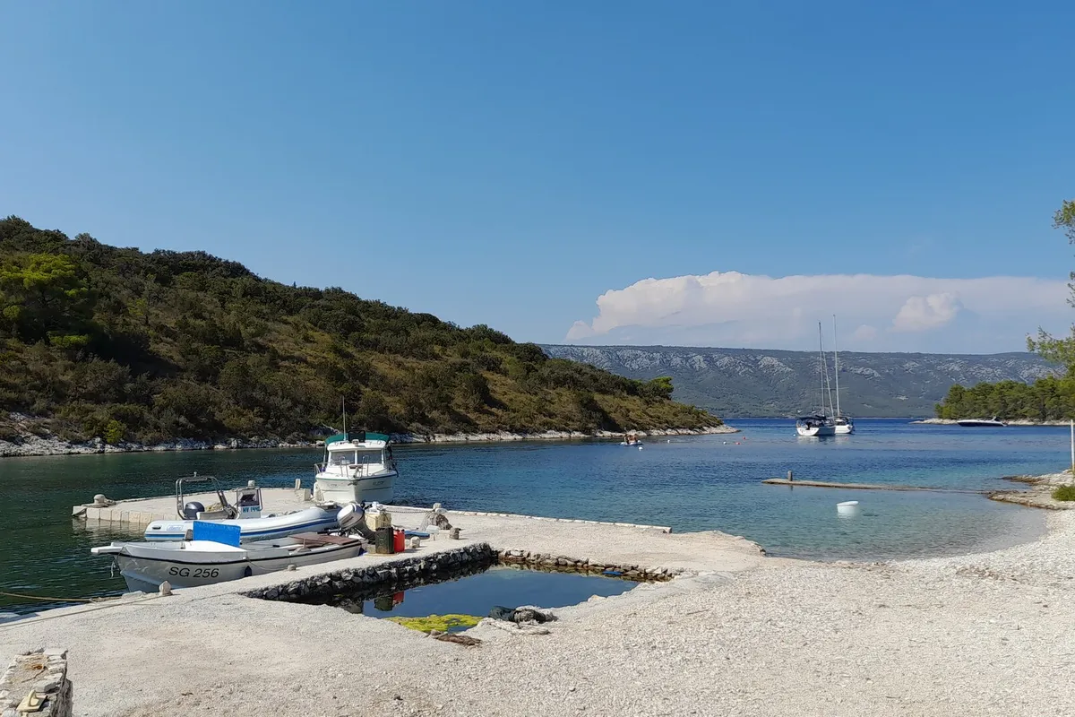 Mostir bay on Šćedro: small boats at a stone pier by the pebble beach, a yacht moored in the bay and the hills of Hvar across the channel