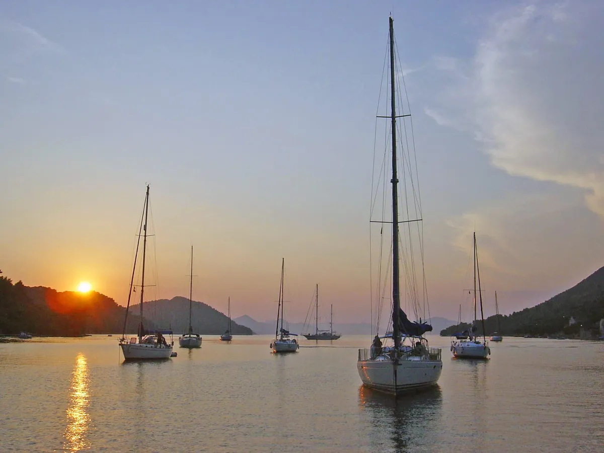 Sailing yachts at anchor in the bay of Šipanska Luka at sunset, with wooded hills on both sides and the sun low over the ridge
