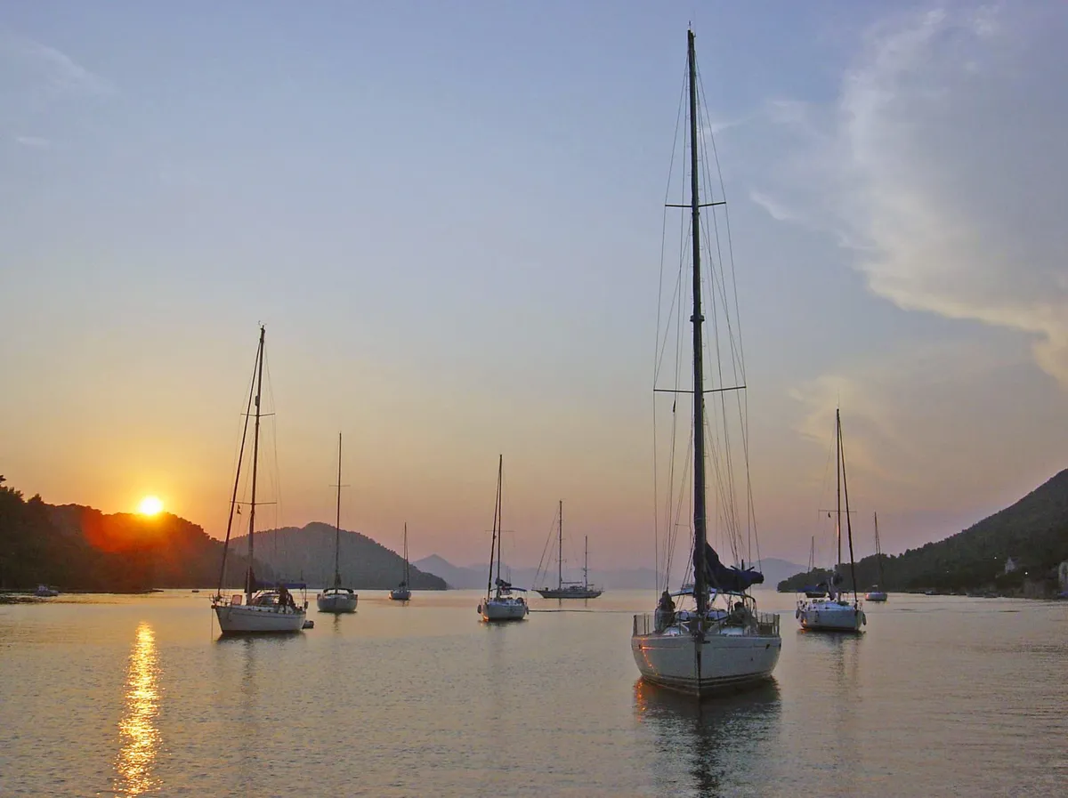 Sailing yachts at anchor in the bay of Šipanska Luka at sunset, with wooded hills on both sides and the sun low over the ridge