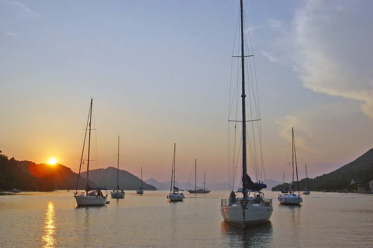 Sailing yachts at anchor in the bay of Šipanska Luka at sunset, with wooded hills on both sides and the sun low over the ridge