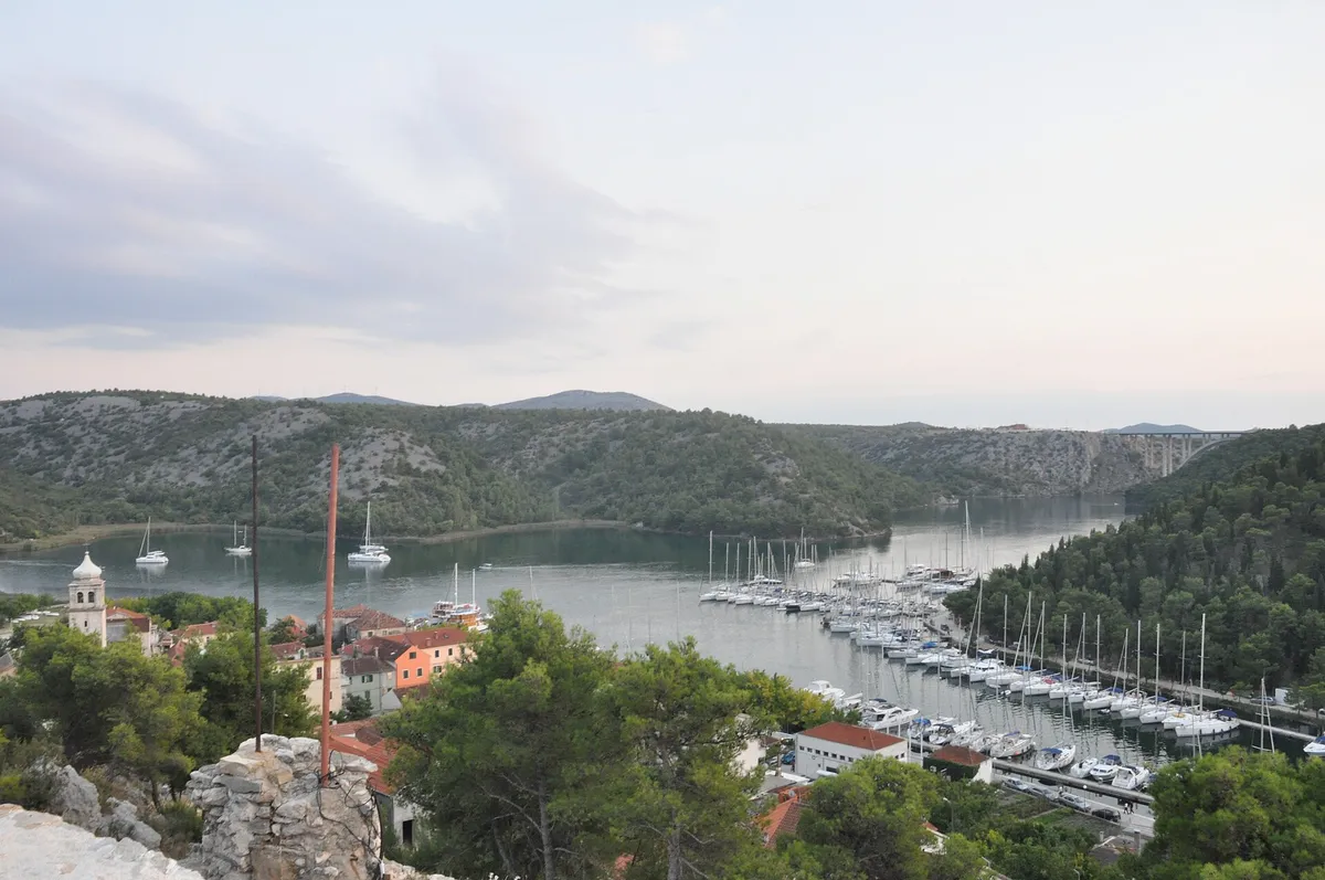ACI Marina Skradin on the Krka river from the hill above the town, yachts on the pontoons and at anchor, the church tower of Skradin at the left and the Krka bridge in the distance
