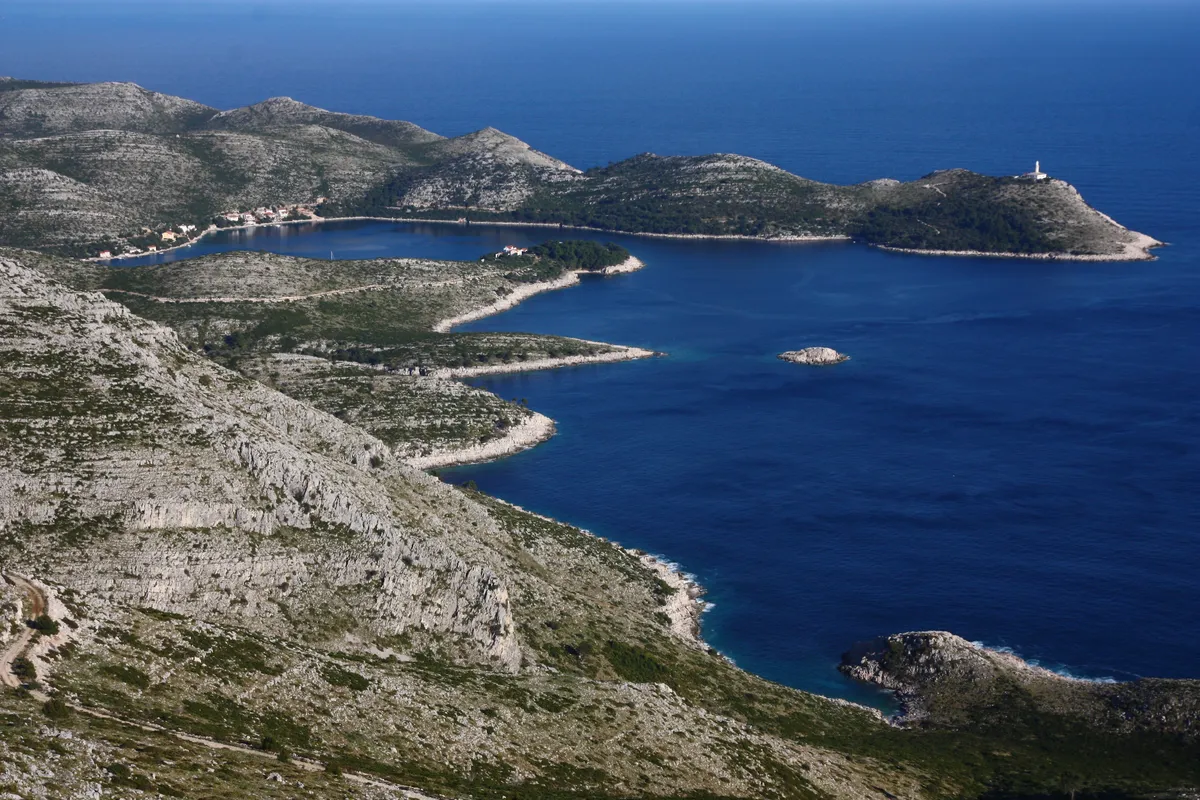 Skrivena Luka on the south coast of Lastovo from the hills above, a long blue bay between bare stony slopes, with the Struga lighthouse on the headland
