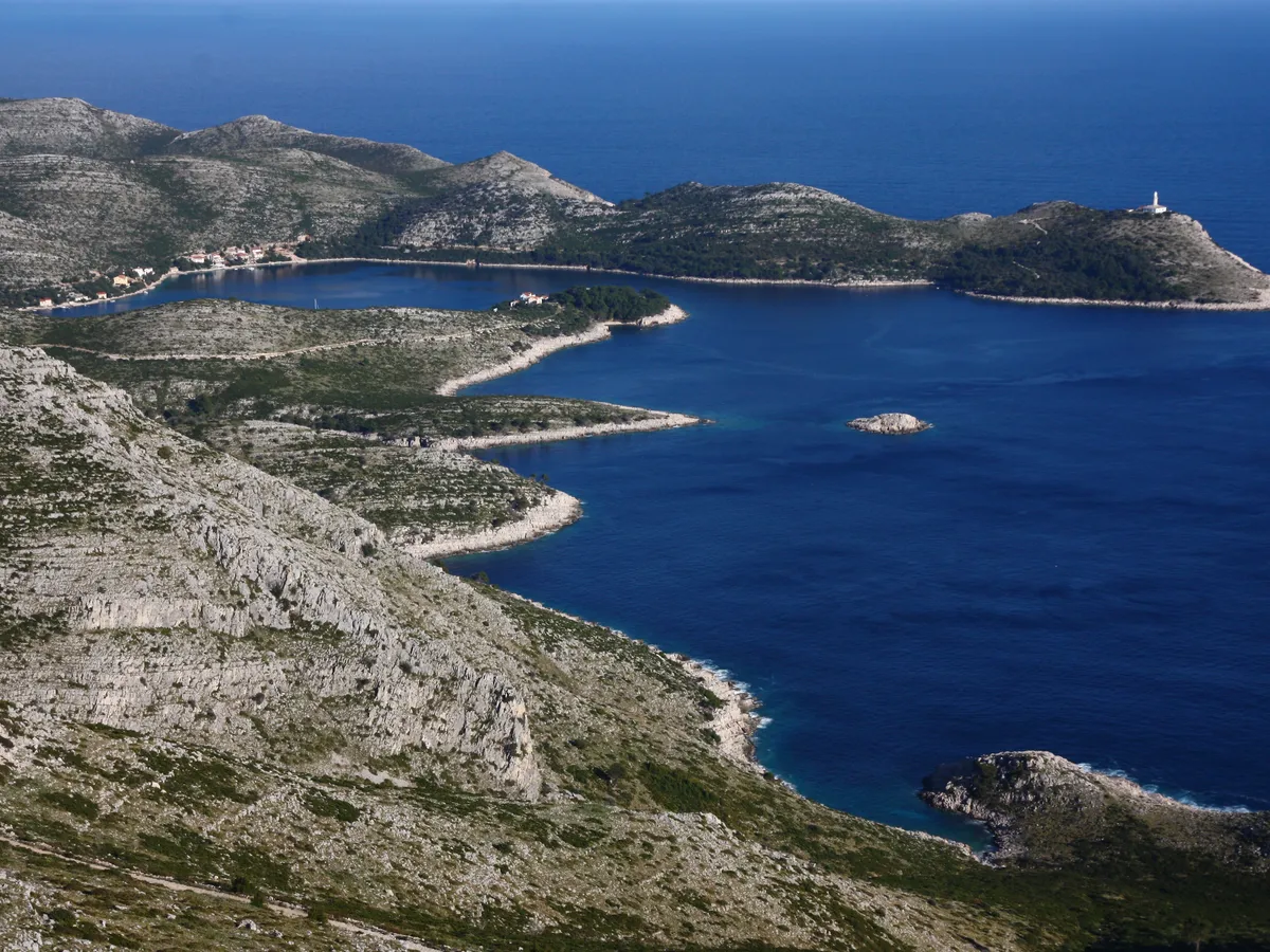 Skrivena Luka on the south coast of Lastovo from the hills above, a long blue bay between bare stony slopes, with the Struga lighthouse on the headland
