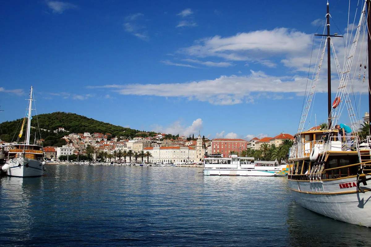 Split's city harbour seen from the water, with a gulet and an excursion boat in front of the Riva and Marjan hill behind.