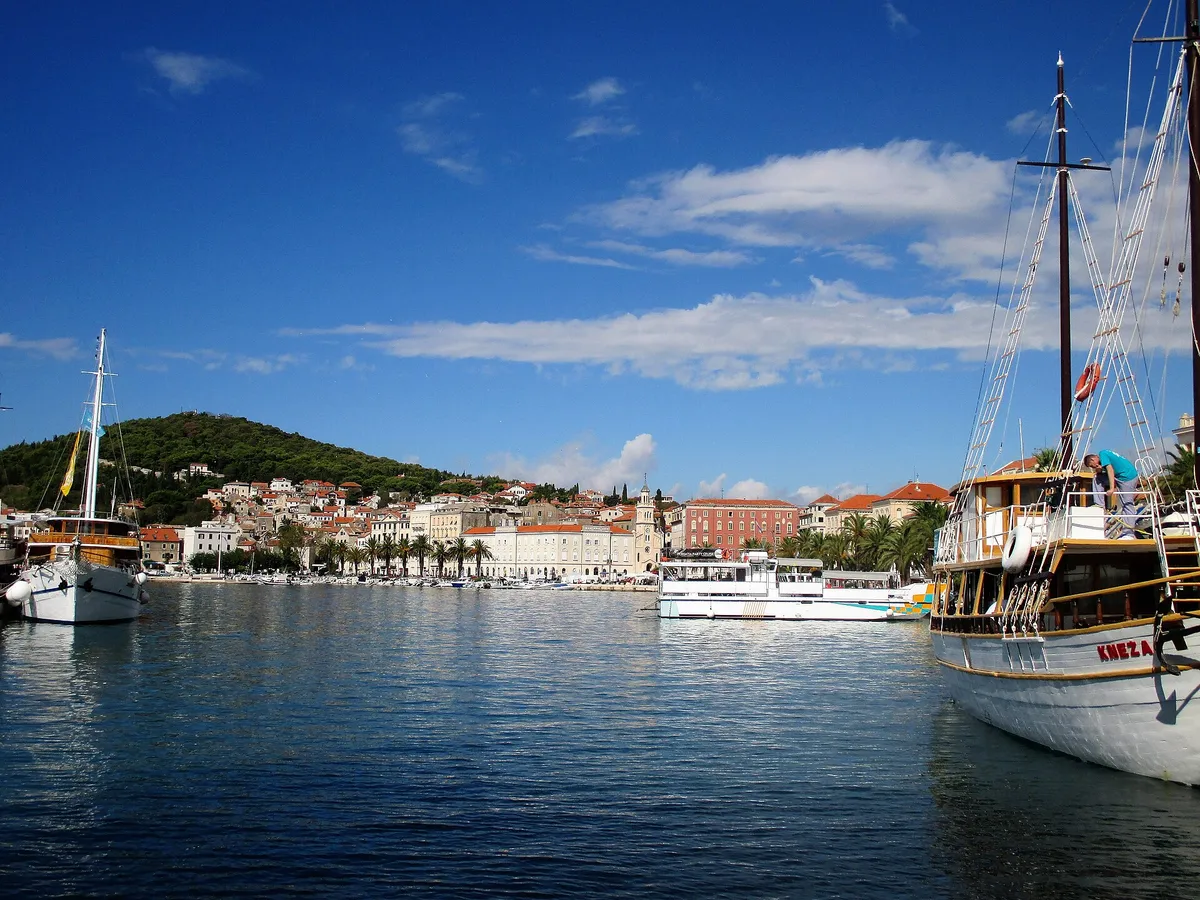 Split's city harbour seen from the water, with a gulet and an excursion boat in front of the Riva and Marjan hill behind.