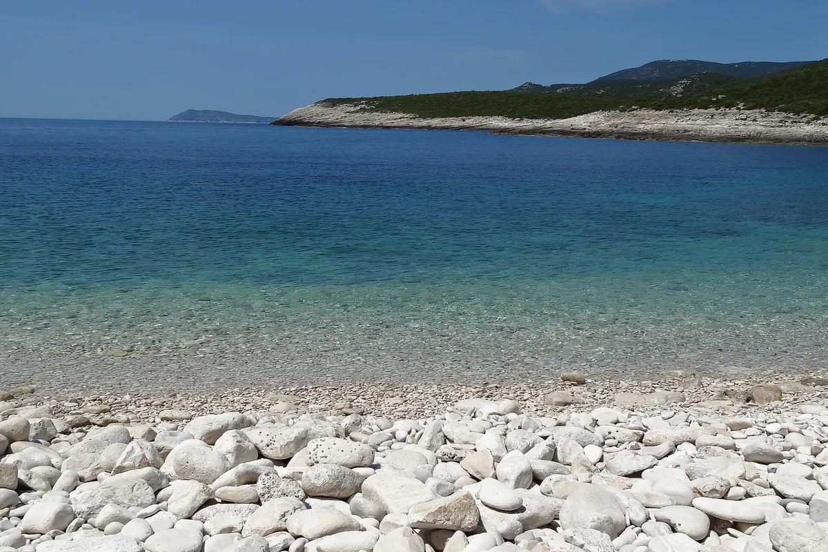 The white pebble beach of Srebrna bay with clear shallow water, the low rock slabs of the headland and open sea beyond