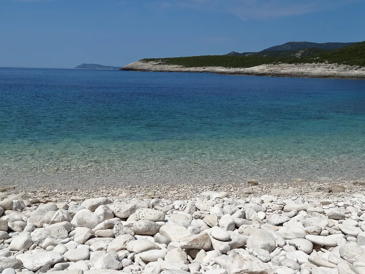 The white pebble beach of Srebrna bay with clear shallow water, the low rock slabs of the headland and open sea beyond