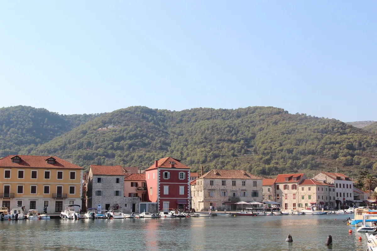 The waterfront of Stari Grad across the inner harbour: old stone houses with red roofs, small boats moored along the quay in front of them and wooded hills behind