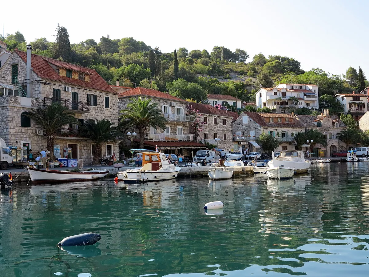 The waterfront of Stomorska on Šolta, with stone houses, palms and small boats on the quay above clear green water.
