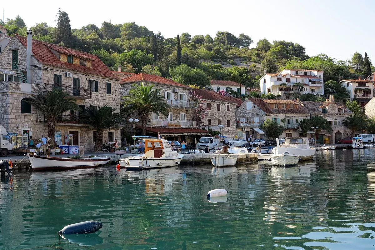 The waterfront of Stomorska on Šolta, with stone houses, palms and small boats on the quay above clear green water.