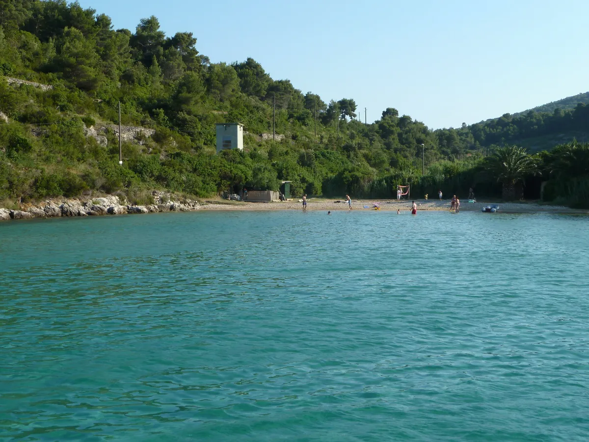 Stončica bay on Vis from the water: turquoise water over sand, the beach at the head of the bay with a few bathers and pine-covered slopes on both sides