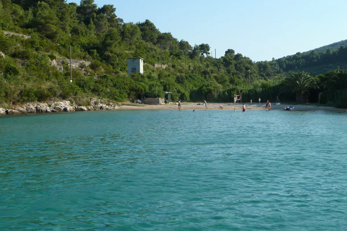 Stončica bay on Vis from the water: turquoise water over sand, the beach at the head of the bay with a few bathers and pine-covered slopes on both sides