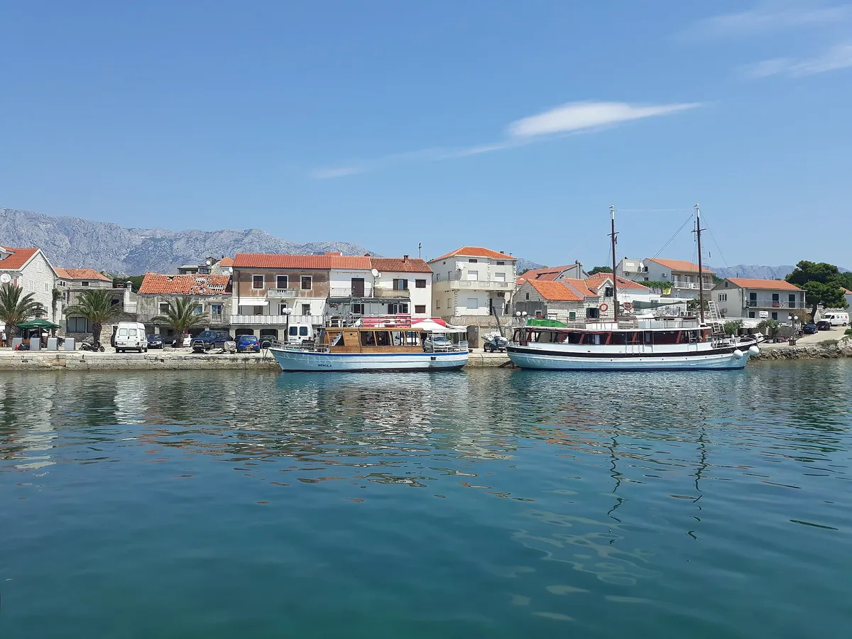 Sućuraj harbour on Hvar from across the water, with an excursion boat and a gulet moored at the stone quay, red-roofed houses behind and the Biokovo mountains on the mainland