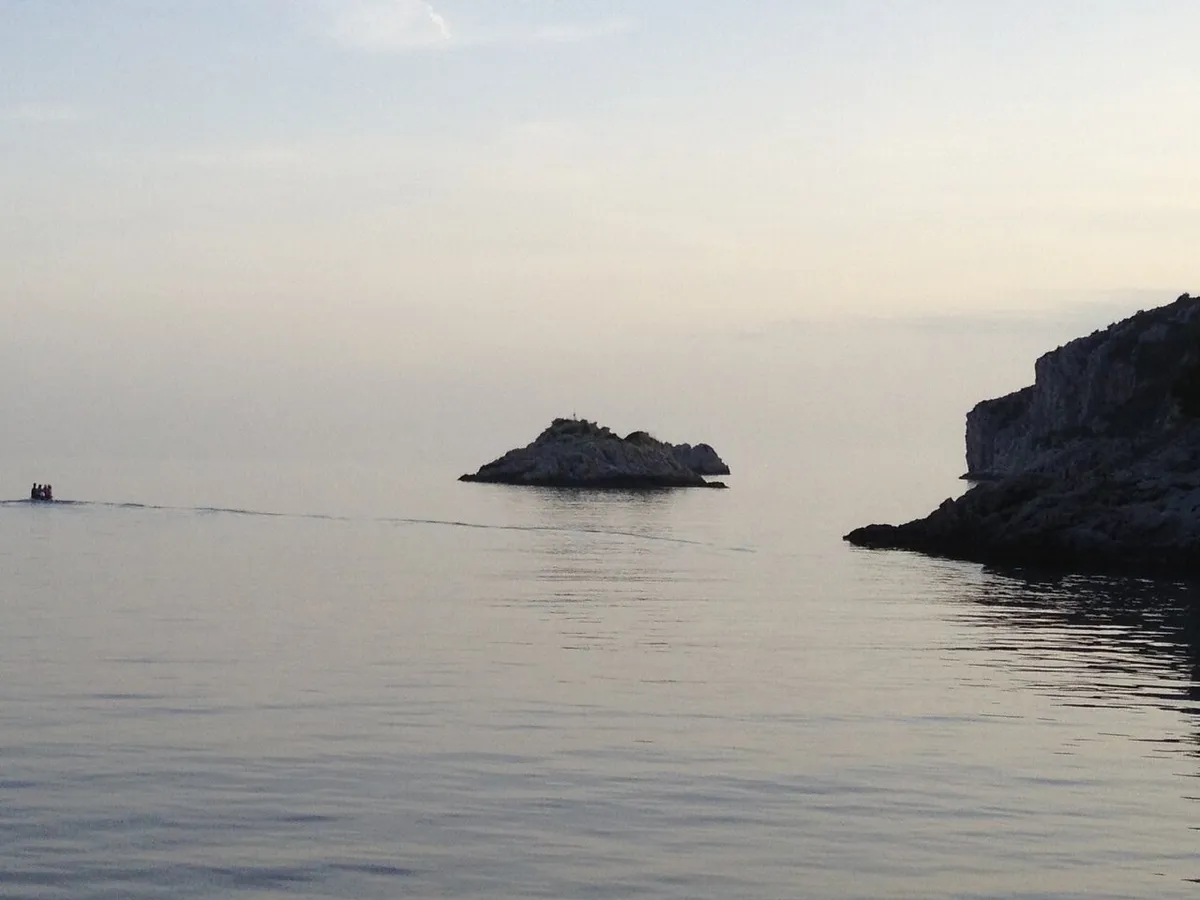 The mouth of Tatinja bay on Šolta at dusk: the Veli Školj rock in a flat calm sea, a small boat heading out and the rocky headland on the right