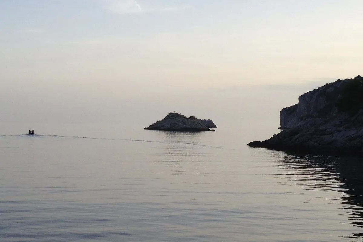 The mouth of Tatinja bay on Šolta at dusk: the Veli Školj rock in a flat calm sea, a small boat heading out and the rocky headland on the right