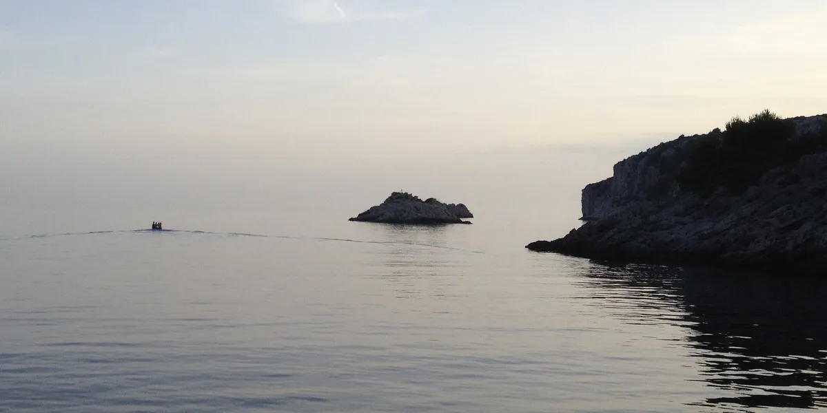 The mouth of Tatinja bay on Šolta at dusk: the Veli Školj rock in a flat calm sea, a small boat heading out and the rocky headland on the right