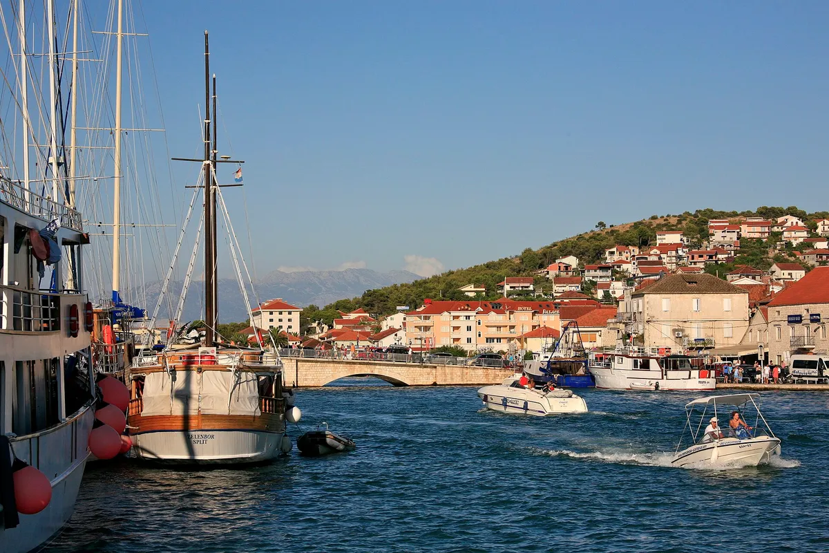Gulets and motorboats in the narrow channel at Trogir, with the old bridge to Čiovo and the Čiovo hillside behind.