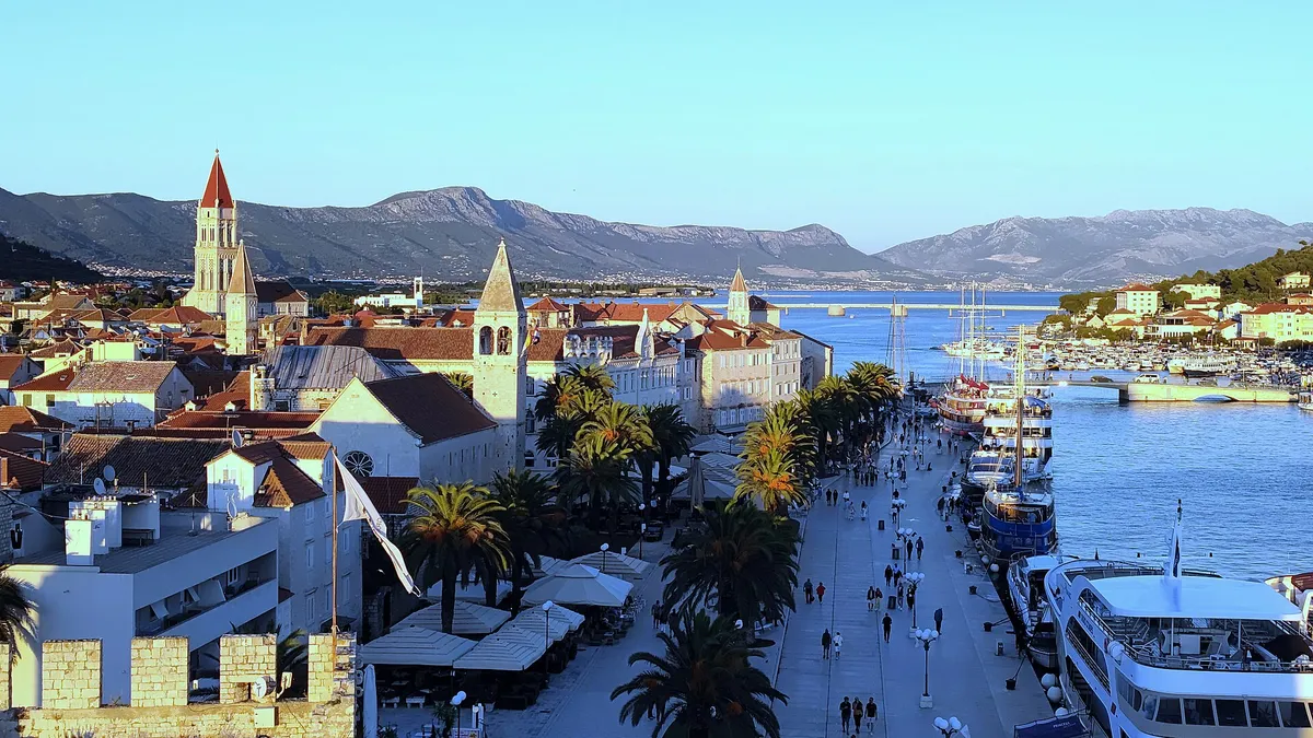 Trogir's Riva from the Kamerlengo fortress, with palms, excursion boats and yachts along the quay and the Čiovo bridge behind.