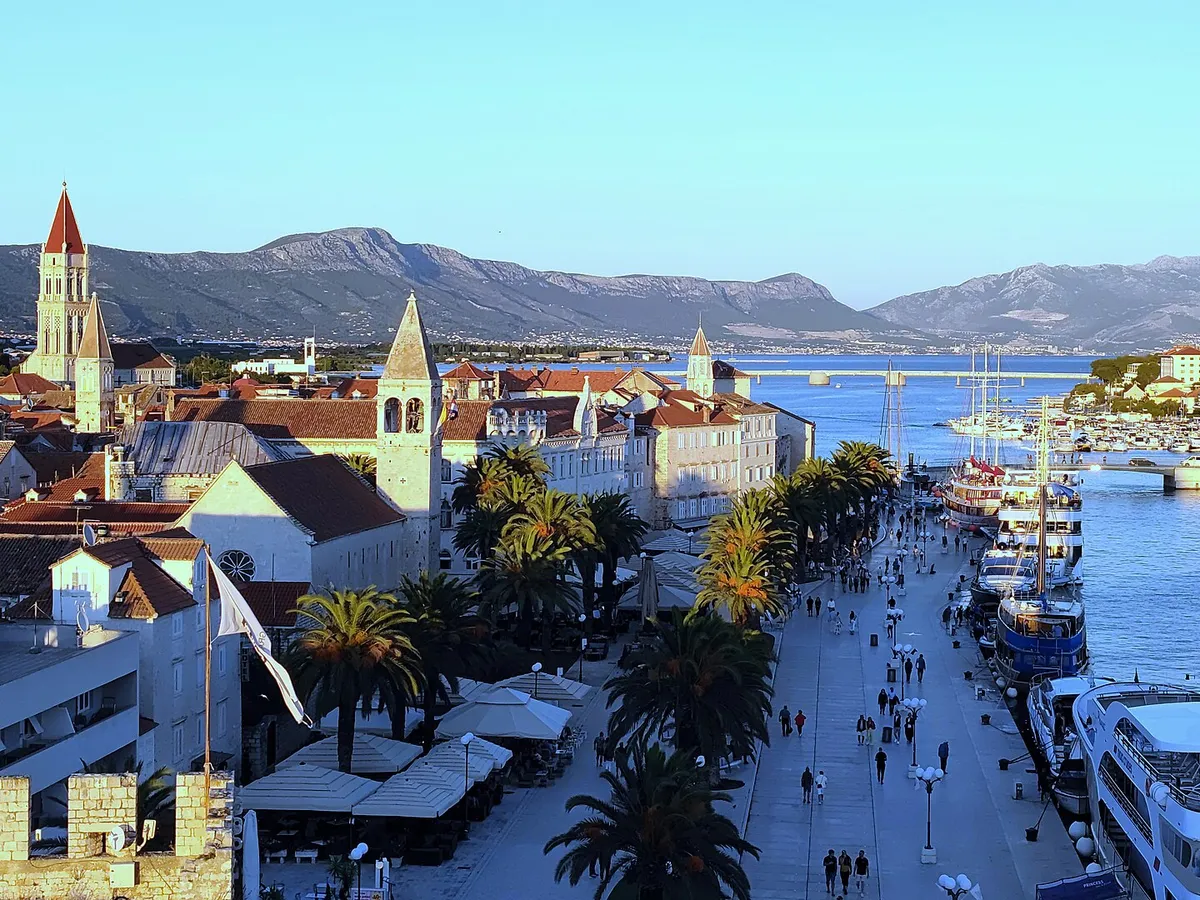 Trogir's Riva from the Kamerlengo fortress, with palms, excursion boats and yachts along the quay and the Čiovo bridge behind.