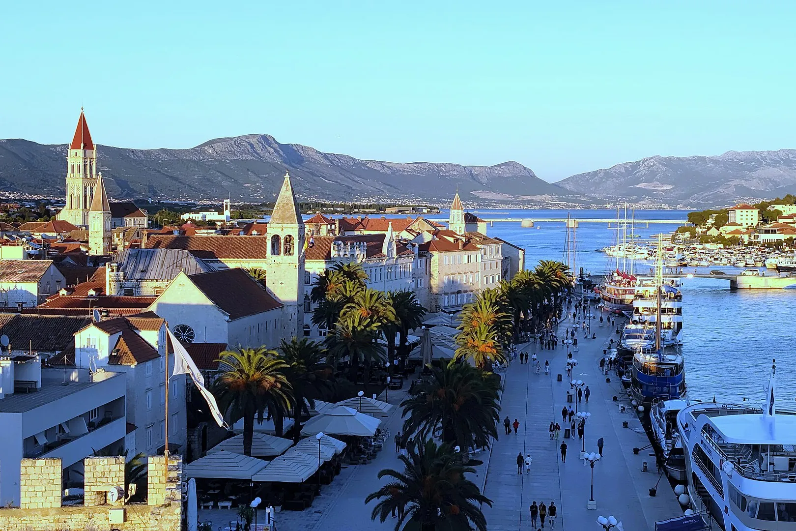 Trogir's Riva from the Kamerlengo fortress, with palms, excursion boats and yachts along the quay and the Čiovo bridge behind.