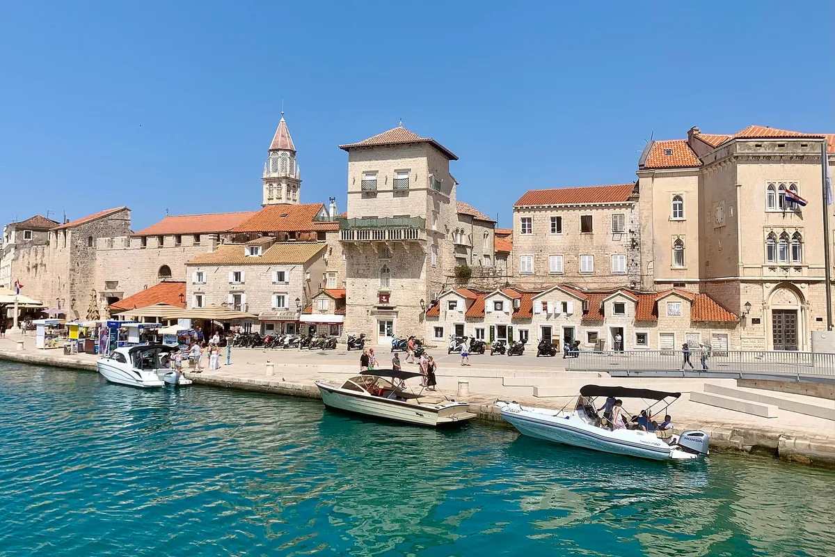 The old town waterfront of Trogir seen from the water, with small motorboats tied along the quay and turquoise water in front.