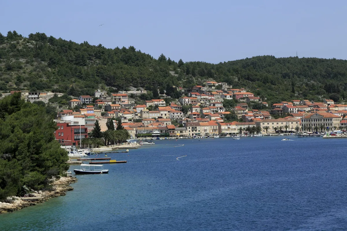 Vela Luka seen from the water, with stone houses and orange roofs along the waterfront under a wooded hill, and a few small boats moored at the shore