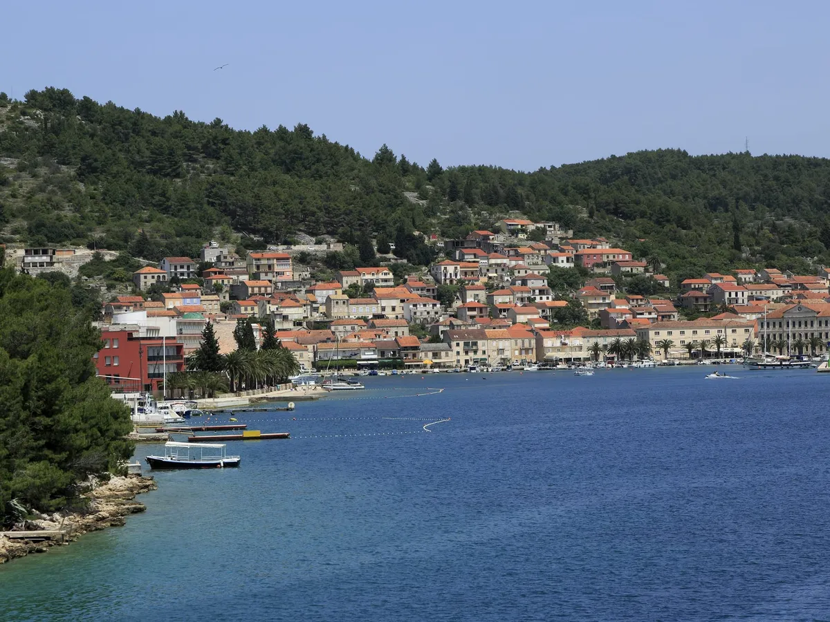 Vela Luka seen from the water, with stone houses and orange roofs along the waterfront under a wooded hill, and a few small boats moored at the shore