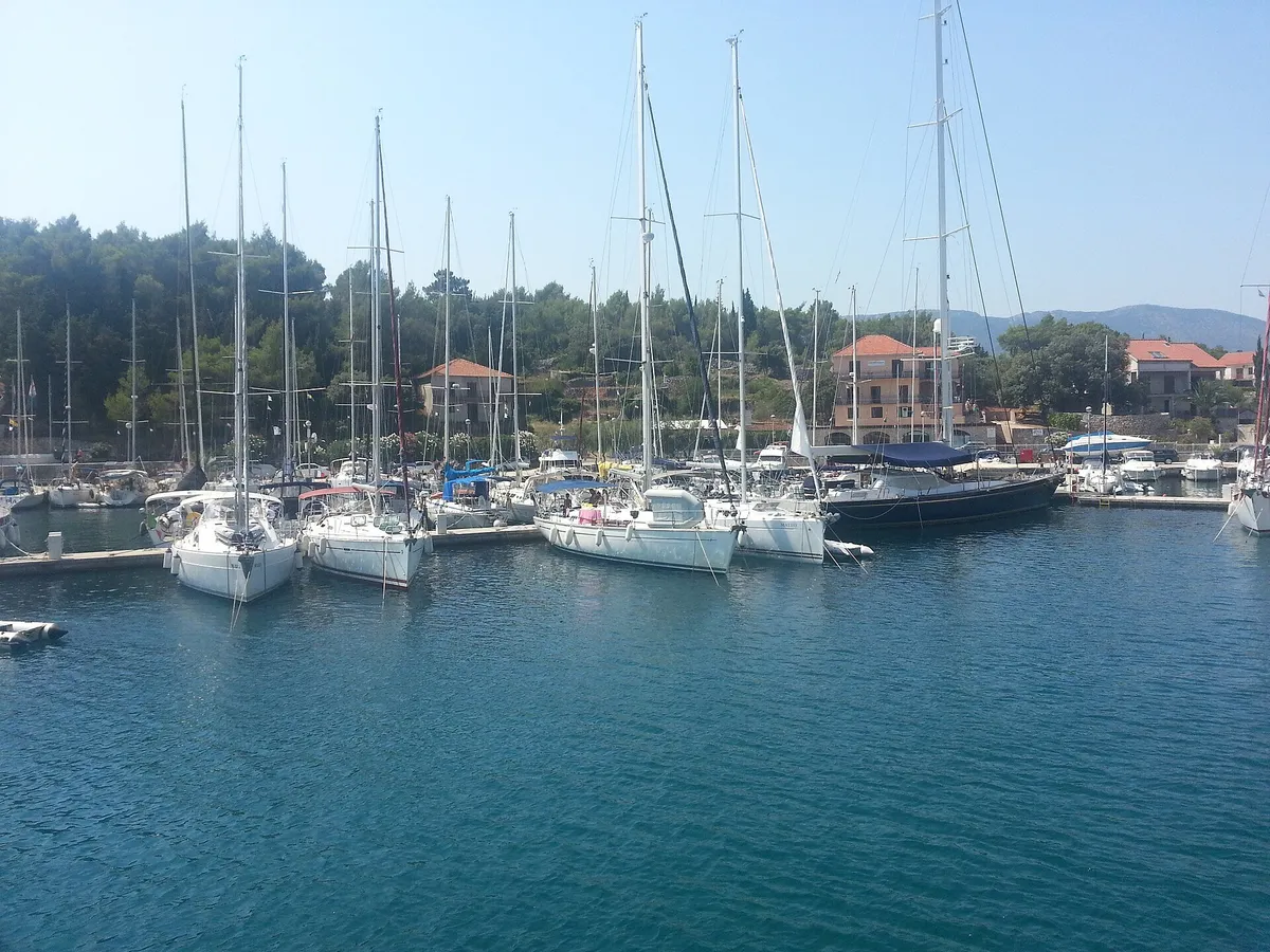 Sailing yachts on the pontoons of ACI Marina Vrboska on Hvar, seen across the water, with pine trees and the marina's buildings behind