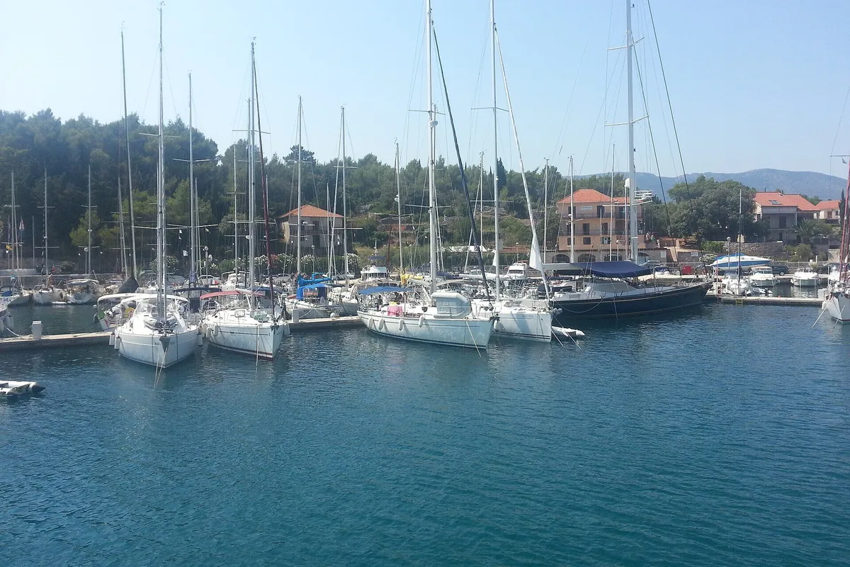 Sailing yachts on the pontoons of ACI Marina Vrboska on Hvar, seen across the water, with pine trees and the marina's buildings behind