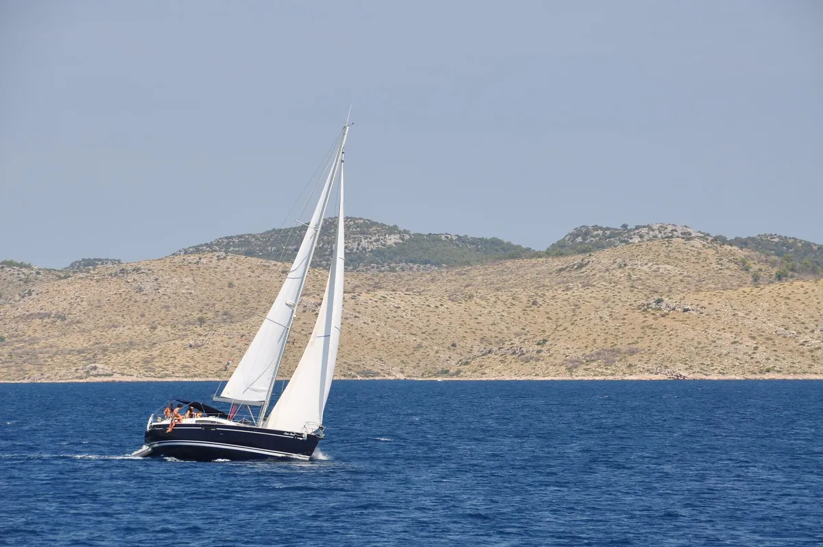 Sailing yacht under full main and genoa on deep blue water, bare Kornati hills behind