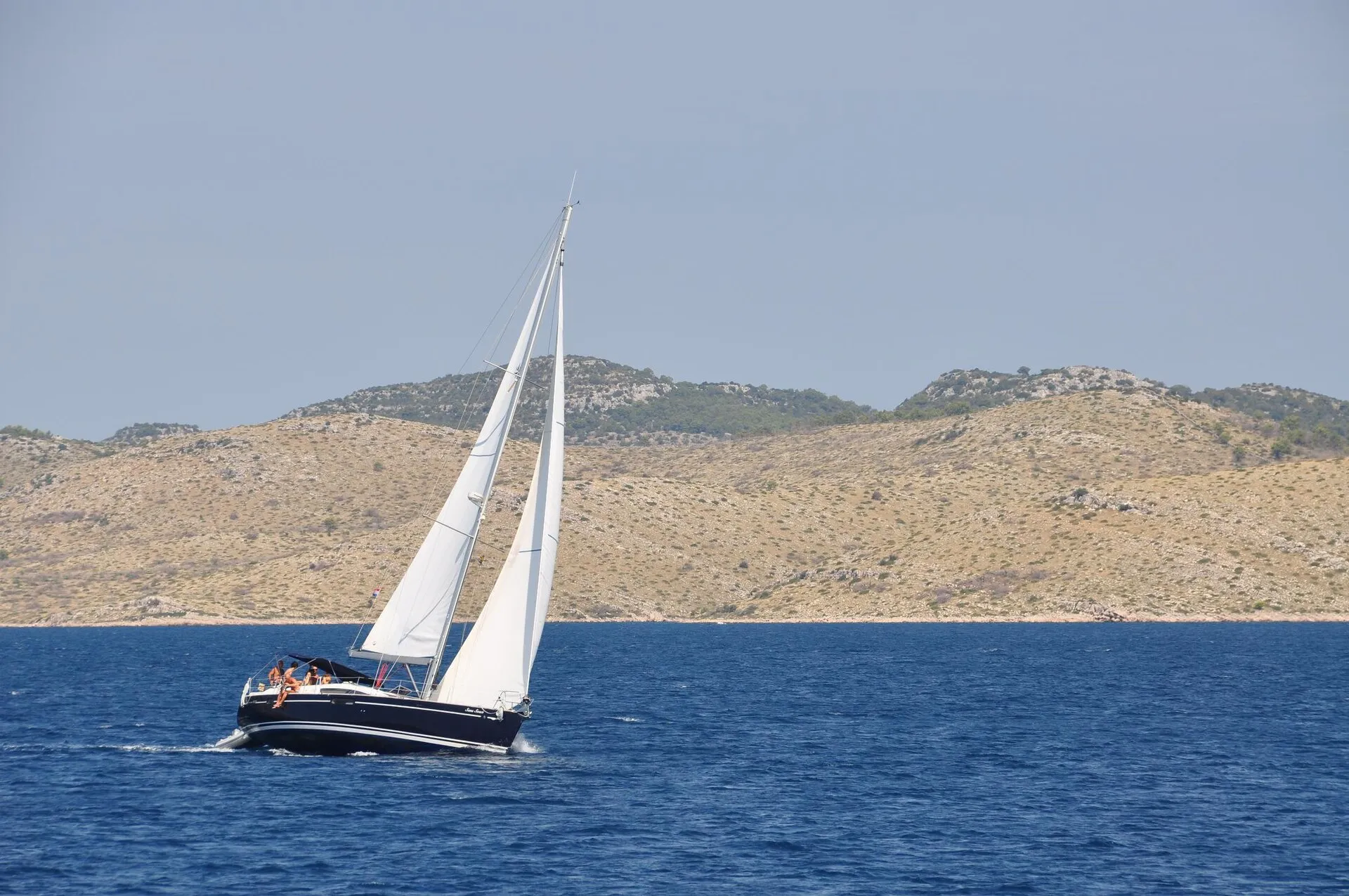 Sailing yacht under full main and genoa on deep blue water, bare Kornati hills behind