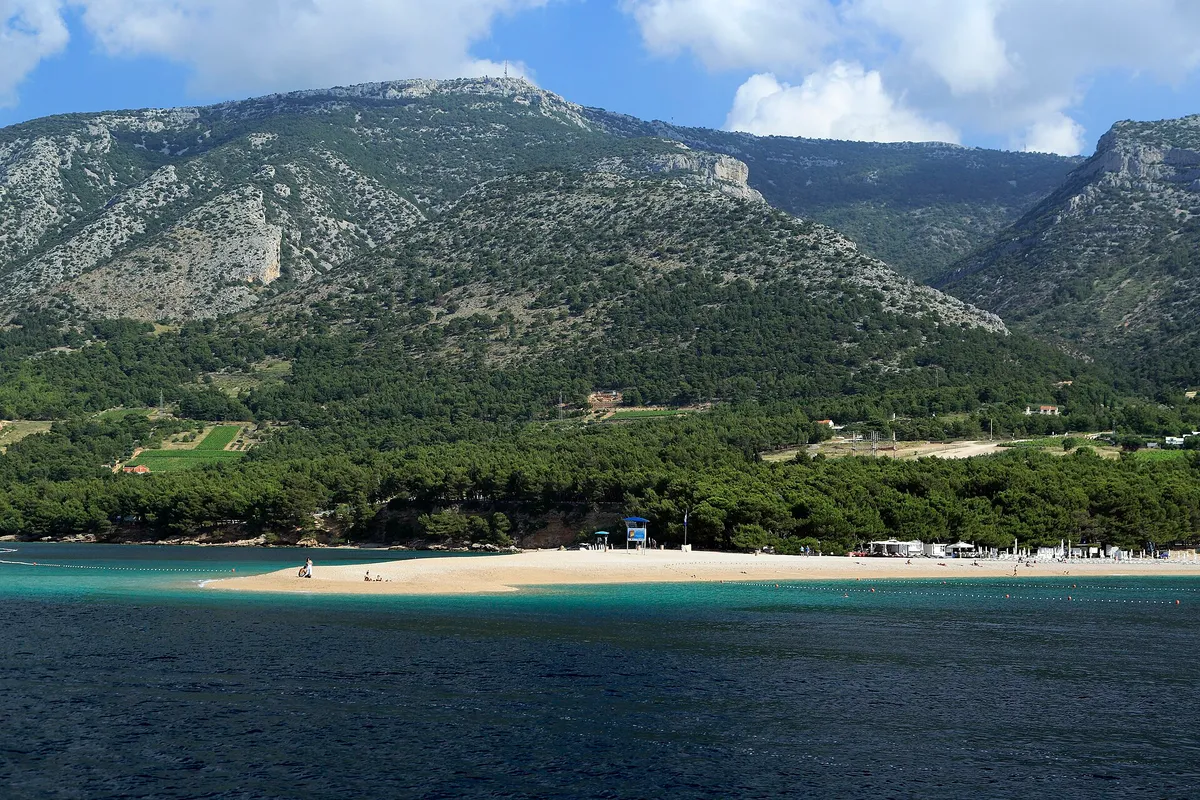 The Zlatni Rat beach spit seen from the sea, with pine woods and Vidova Gora rising behind.